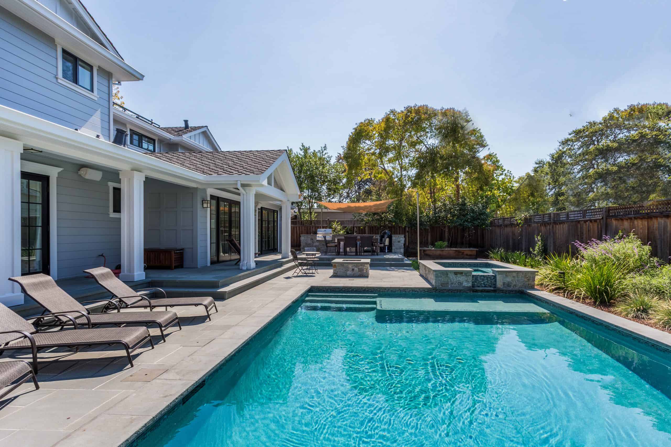 A modern backyard with a swimming pool, lounge chairs, shaded patio, and tall trees in the background. The house has a grey exterior, and there's a convenient spot for propane delivery to fuel your outdoor grill.
