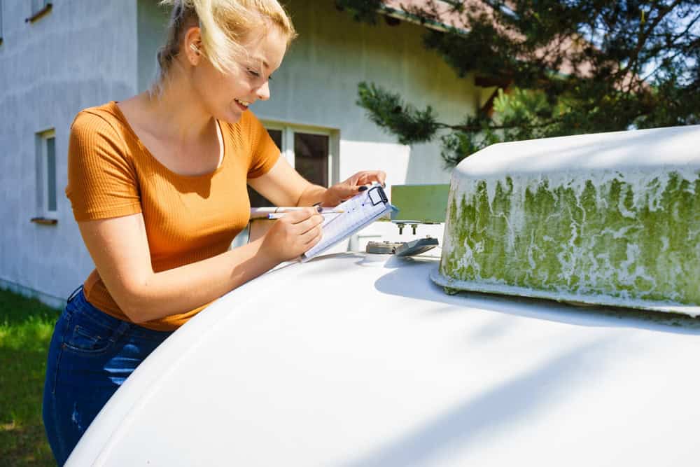 A woman with blond hair in an orange shirt examines and writes on a clipboard outside near a large white propane tank with a green, weathered top.