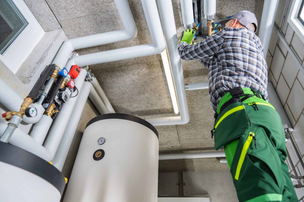 A plumber in green work pants and gloves fixes pipes on a ceiling in a utility room with a large water heater, various pipes, and propane services equipment.