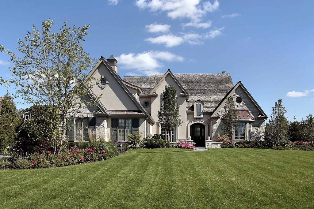 Two-story house with stone and stucco exterior, arched windows, and extensive landscaping including a manicured lawn and flower beds under a blue sky. Convenient propane services available with seamless fuel tank installation ensure comfort year-round.