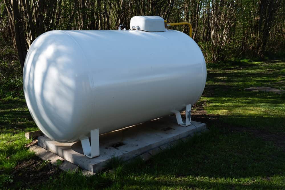 A large white propane tank sits on a concrete pad in a grassy area surrounded by trees, ready for its next propane refill.