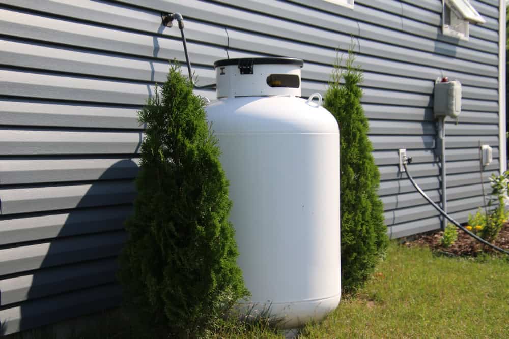 A white propane tank, crucial for fuel tank installation, is placed outside of a grey house, flanked by two small evergreen bushes. The tank is connected to the house by a hose.