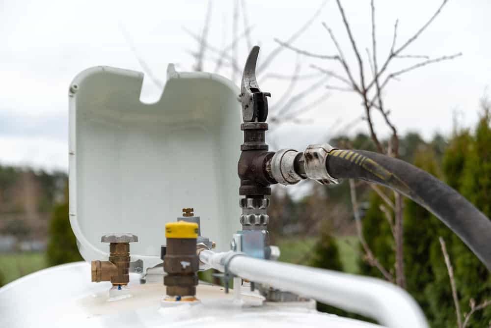 Close-up view of an outdoor propane tank with an open white cap, showing pipes, valves, and a connected hose, surrounded by trees.