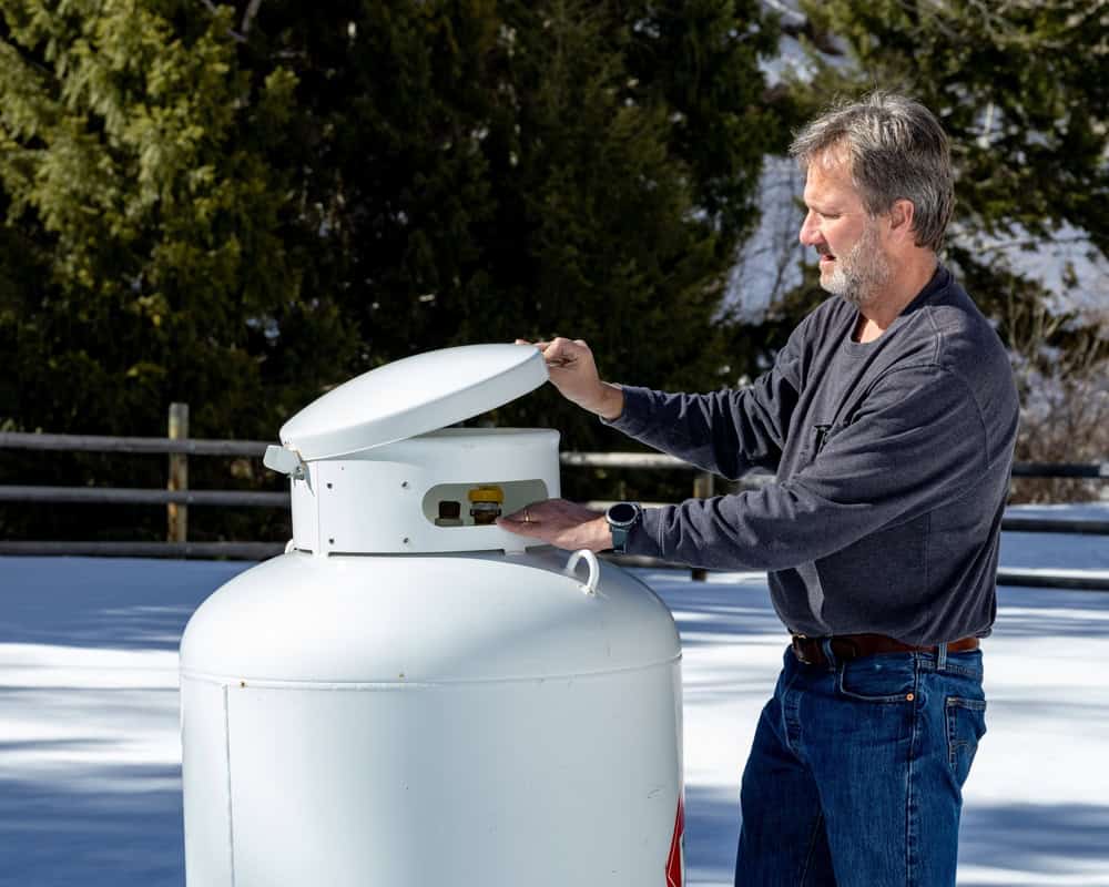 A man in a gray long-sleeve shirt and jeans opens the lid of a large white propane tank outdoors on a snowy day, preparing for propane delivery with trees in the background.