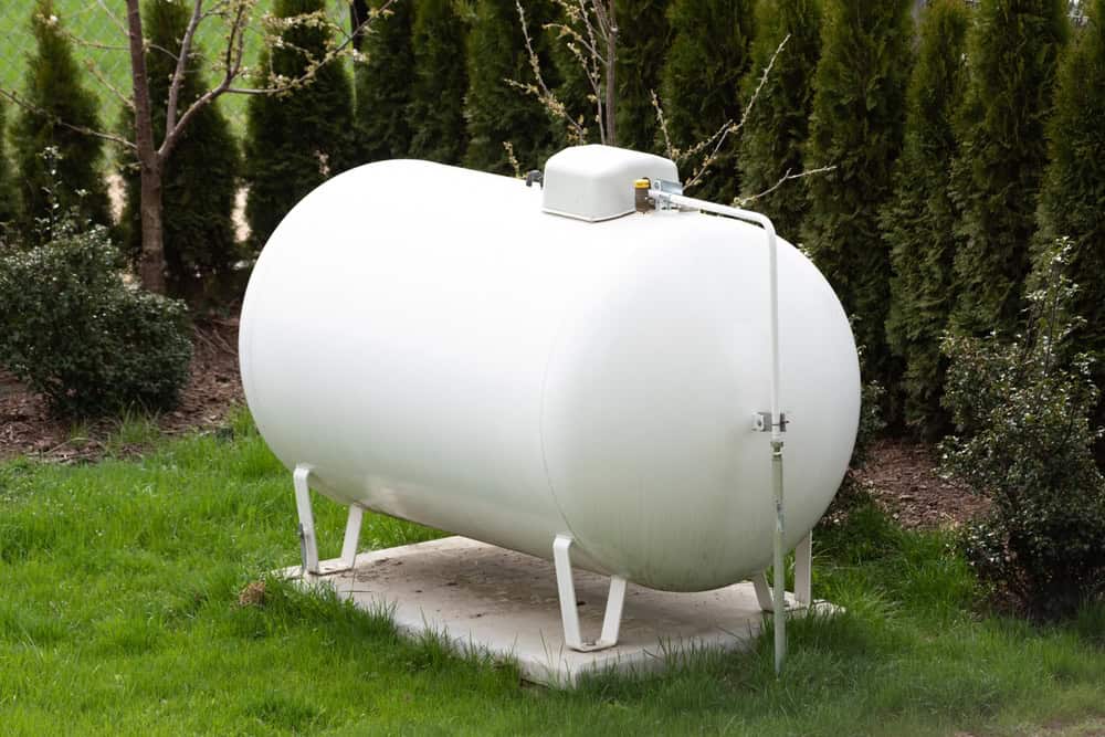 A white propane tank situated on a concrete slab surrounded by grass and trees, with pipes attached to the top, showcases an impeccable example of seamless fuel tank installation.