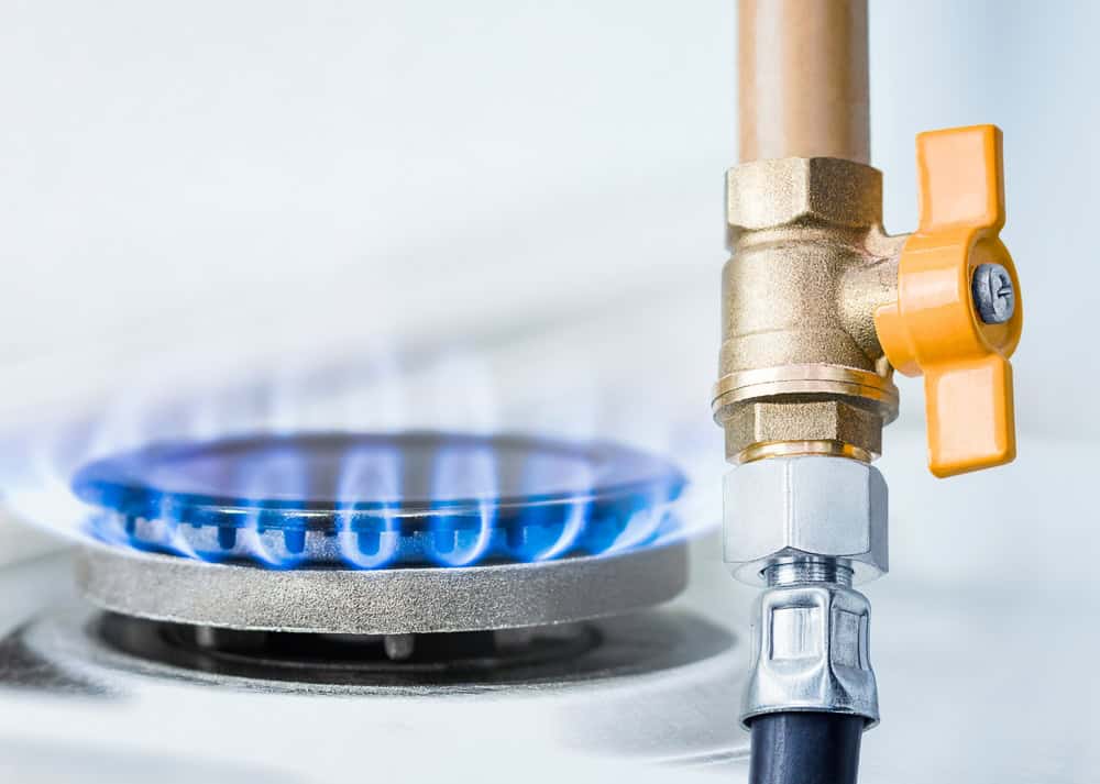 Close-up of a lit gas stove burner with blue flames and a brass valve with an orange handle connected to a gas pipe, showcasing the precision of propane delivery.