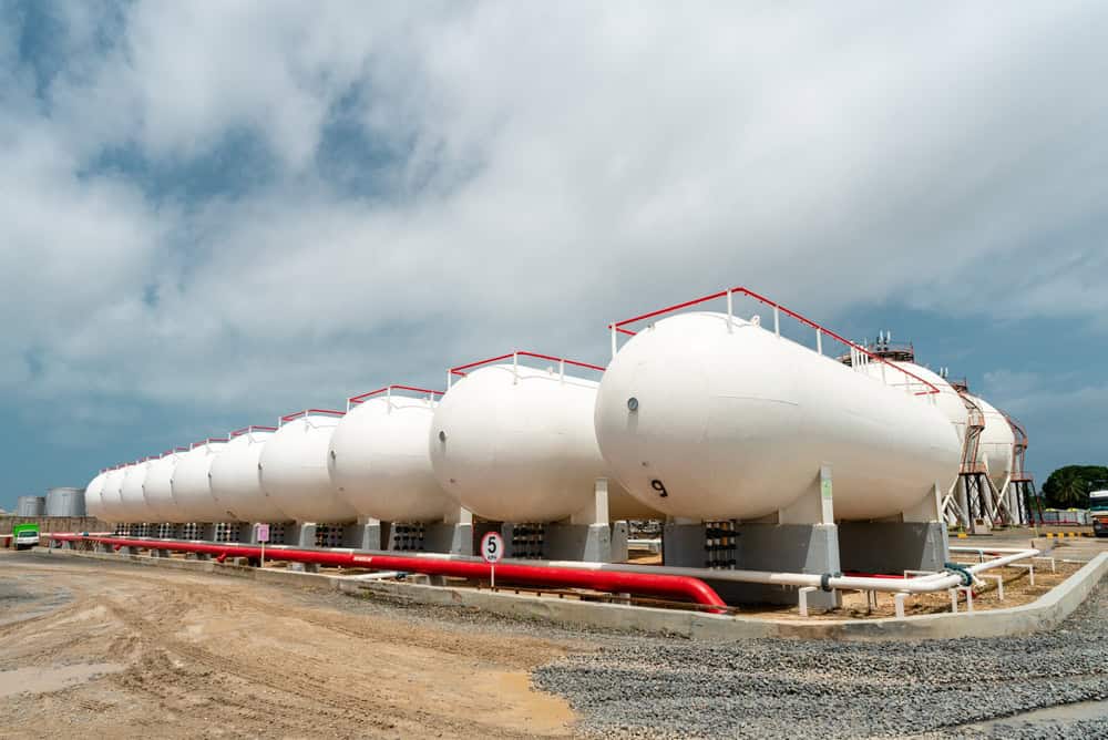 A row of large, white cylindrical storage tanks stands on a gravel and dirt area, awaiting the next propane delivery under a blue sky with scattered clouds in the background.