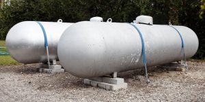 Two large horizontal silver propane tanks, installed with Fuel tank installation services, are secured with blue straps and placed on concrete blocks outdoors, surrounded by trees.