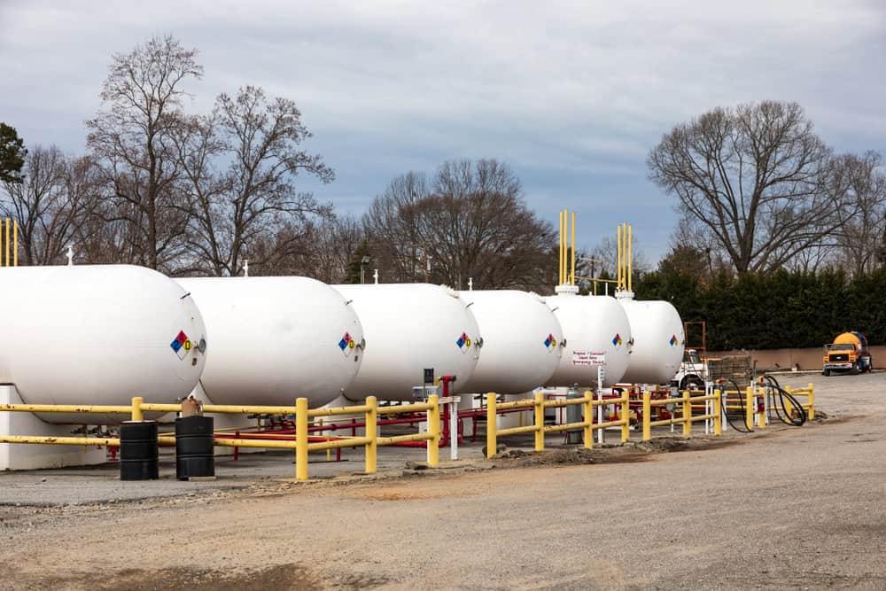 White propane storage tanks with hazard symbols, surrounded by yellow railings, stand outdoors on a cloudy day. A nearby truck suggests ongoing propane delivery services. Trees are visible in the background.