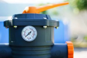 Close-up of a black water filter housing with a pressure gauge and an orange valve handle on top, outdoors in Suffolk County, NY—perfect for properties using propane services Suffolk County residents trust. Blurred background highlights the setup.