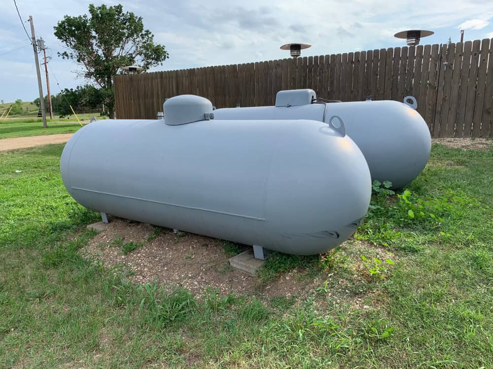 Two large gray propane tanks, awaiting a propane refill, are placed on grassy ground near a wooden fence, with a clear blue sky in the background.