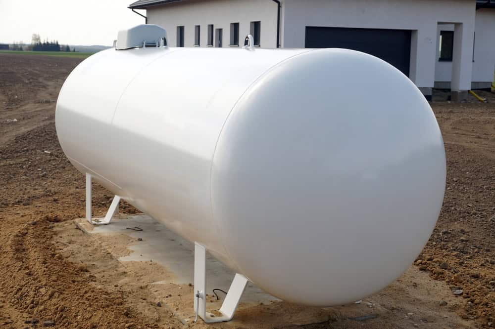 An outdoor white propane tank stands on a sandy yard area next to a house with a garage, ready for convenient propane refill or delivery.