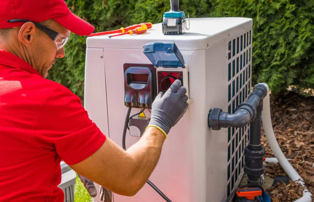 A technician in a red shirt and cap adjusts settings on an outdoor HVAC unit beside a building, ensuring everything runs smoothly for the upcoming propane delivery.