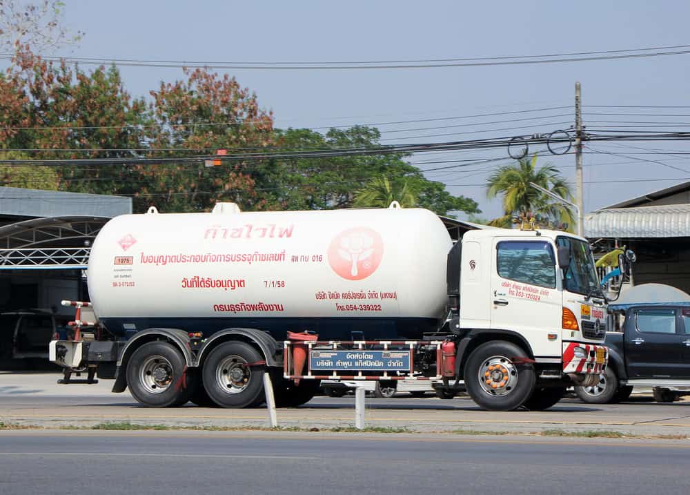 A white tanker truck with red text parked on the side of a road, near a building and other vehicles. Trees and power lines are visible in the background, signifying efficient propane delivery in progress.