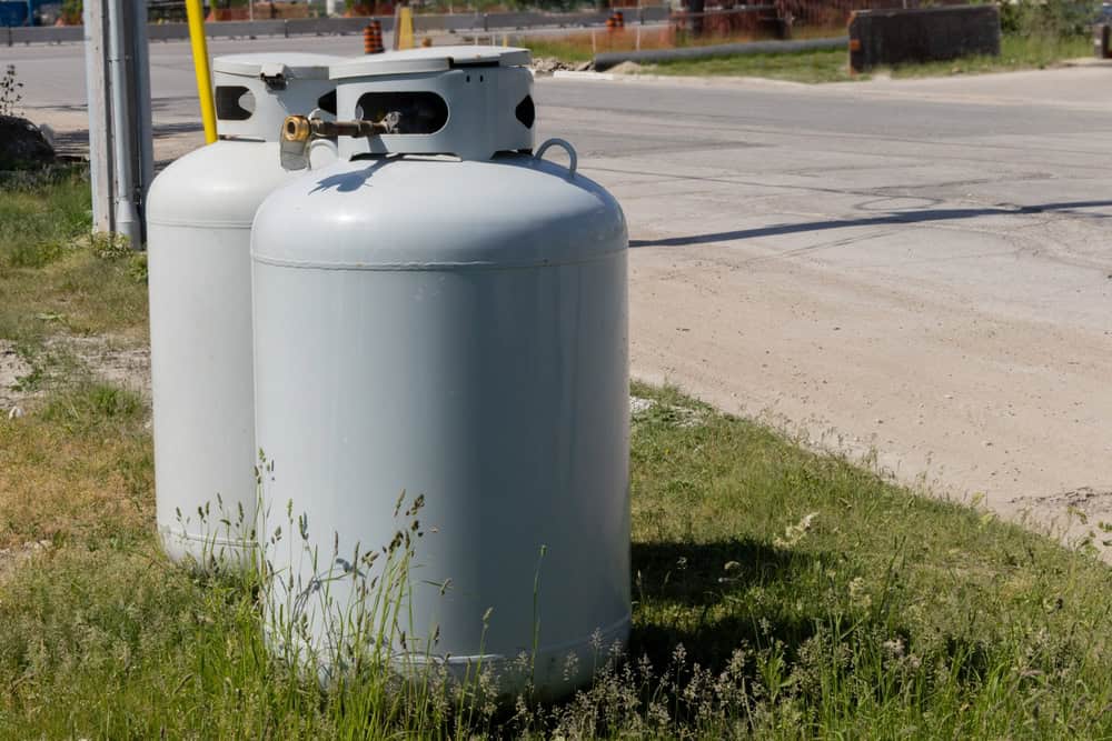 Two large metal propane tanks, ideal for an emergency propane delivery in Suffolk County, are positioned on grass near a road. The tanks are cylindrical with rounded tops and visible valves and handles. The background shows a street with some construction barriers and industrial elements.
