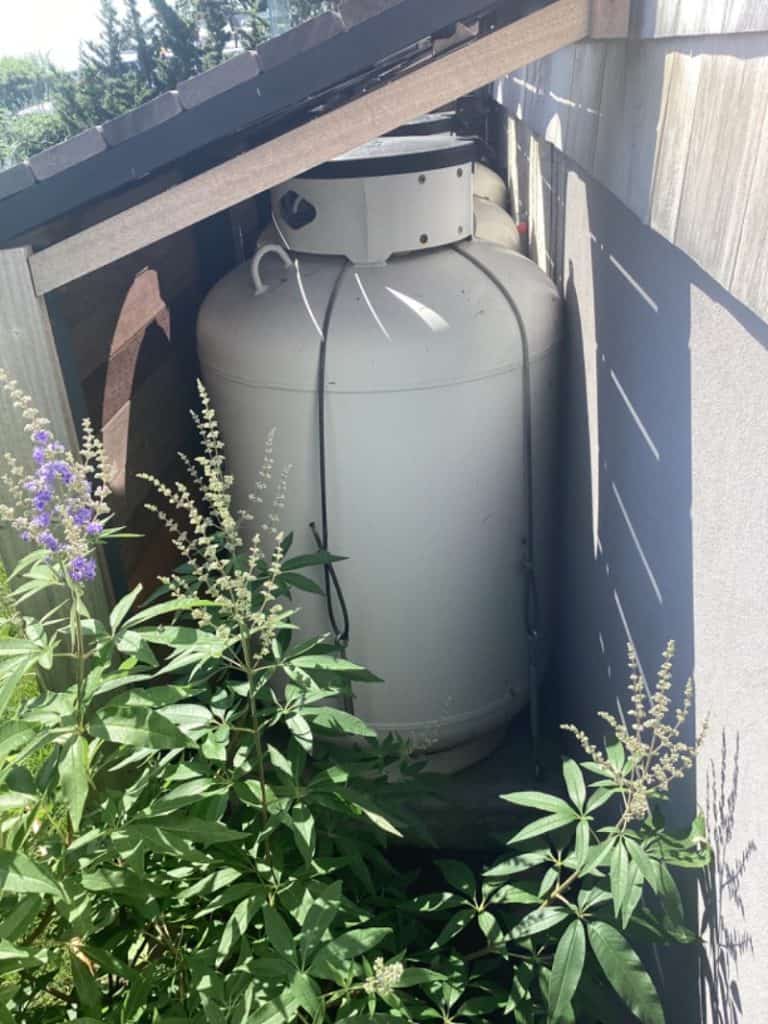 A large white propane tank stands in a narrow outdoor space between a building and a fence, surrounded by green plants with purple flowers and partially shaded by the roof above, illustrating why propane is ideal for versatile outdoor energy needs.