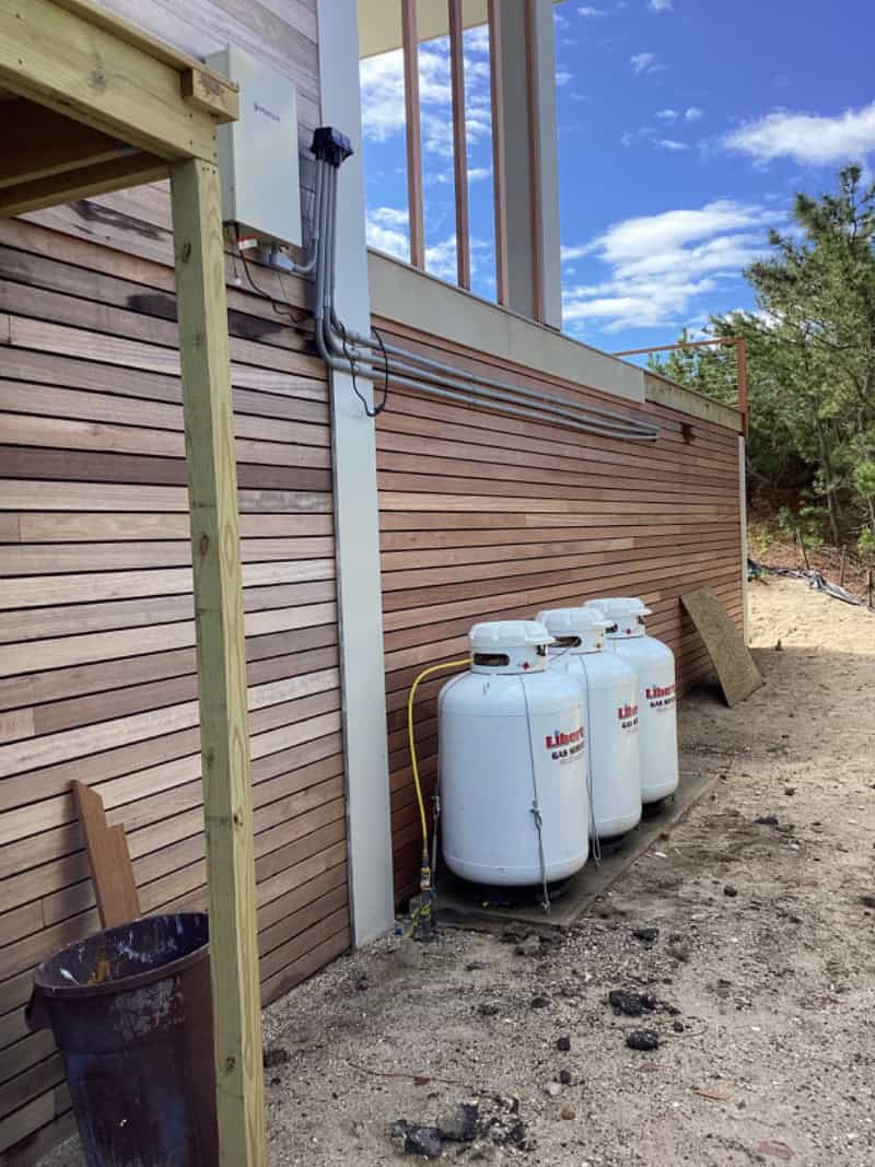 Four large propane tanks are lined up on a concrete pad next to a wooden building with horizontal siding&mdash;an example of why propane is chosen for reliable fuel. Pipes and cables run above the tanks; sand and sparse vegetation surround the area.
