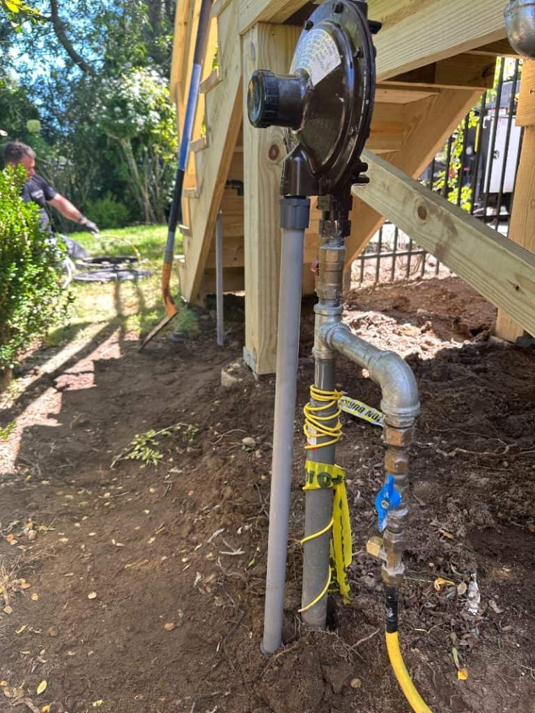 A close-up of a natural gas regulator and pipe installation under a wooden deck, with yellow flexible tubing and valves; highlighting safety, this scene subtly answers why propane is often chosen as someone works near greenery in the background.