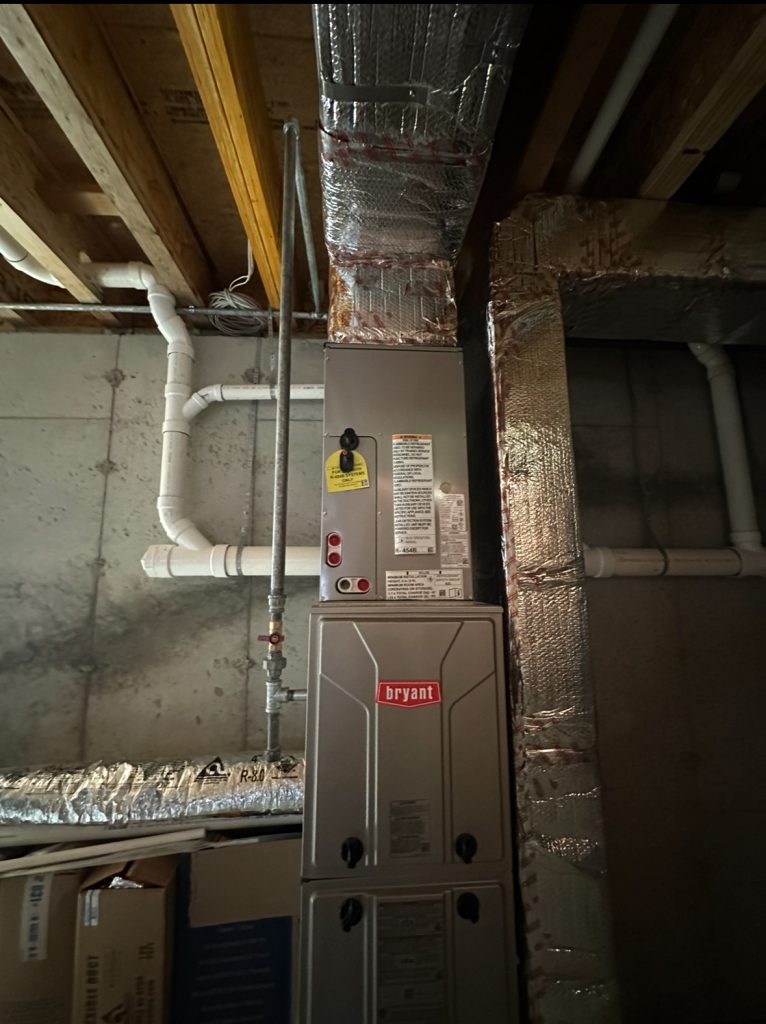 A vertical view of a Bryant furnace unit with vents and pipes attached, installed in a basement with exposed beams and concrete walls. The area is dimly lit, showcasing work done by the local Propane Company.