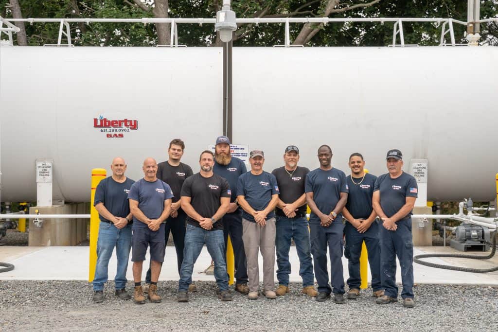 Ten men in navy work shirts and jeans stand together, smiling in front of a large white Liberty Gas tank. Their teamwork reflects Liberty Gas’s commitment to safety and ongoing training, with trees and gravel visible in the background.