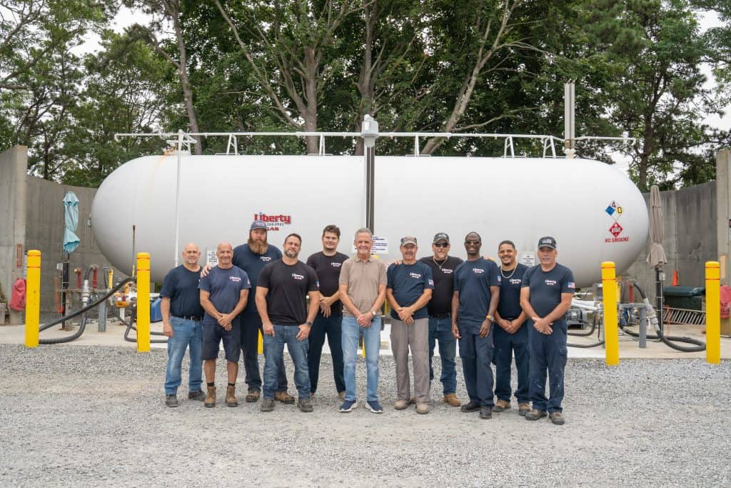 A group of twelve workers in blue uniforms stands smiling in front of a large white propane tank, showcasing their expertise in Propane Service & Installation. They are outdoors on gravel, with safety poles and trees in the background.