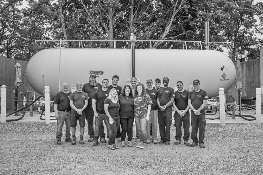 A group of 16 people, mostly men, stand together smiling in front of a large industrial propane tank outdoors, surrounded by trees and gravel. The team, dressed in casual work attire, represents a dedicated crew from Propane Services.