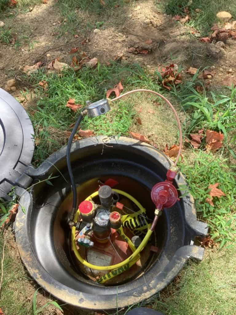 A partially buried propane tank with its lid open, showing valves, a pressure regulator, connected hoses, and green grass with some brown, fallen leaves around it—highlighting why propane is a reliable choice for outdoor energy needs.