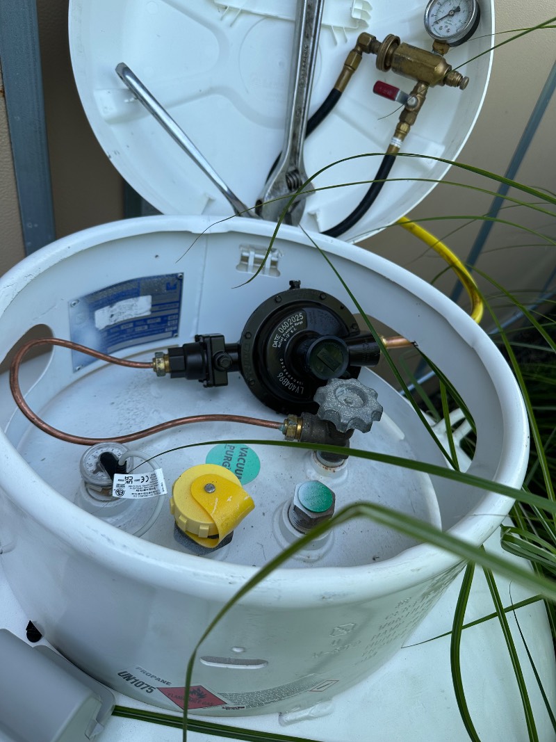 Close-up of an open propane tank with a regulator, hoses, pressure gauge, and various valves; blades of grass partly cover the tank, tools are visible under the open lid—showcasing details essential for reliable Propane Services.