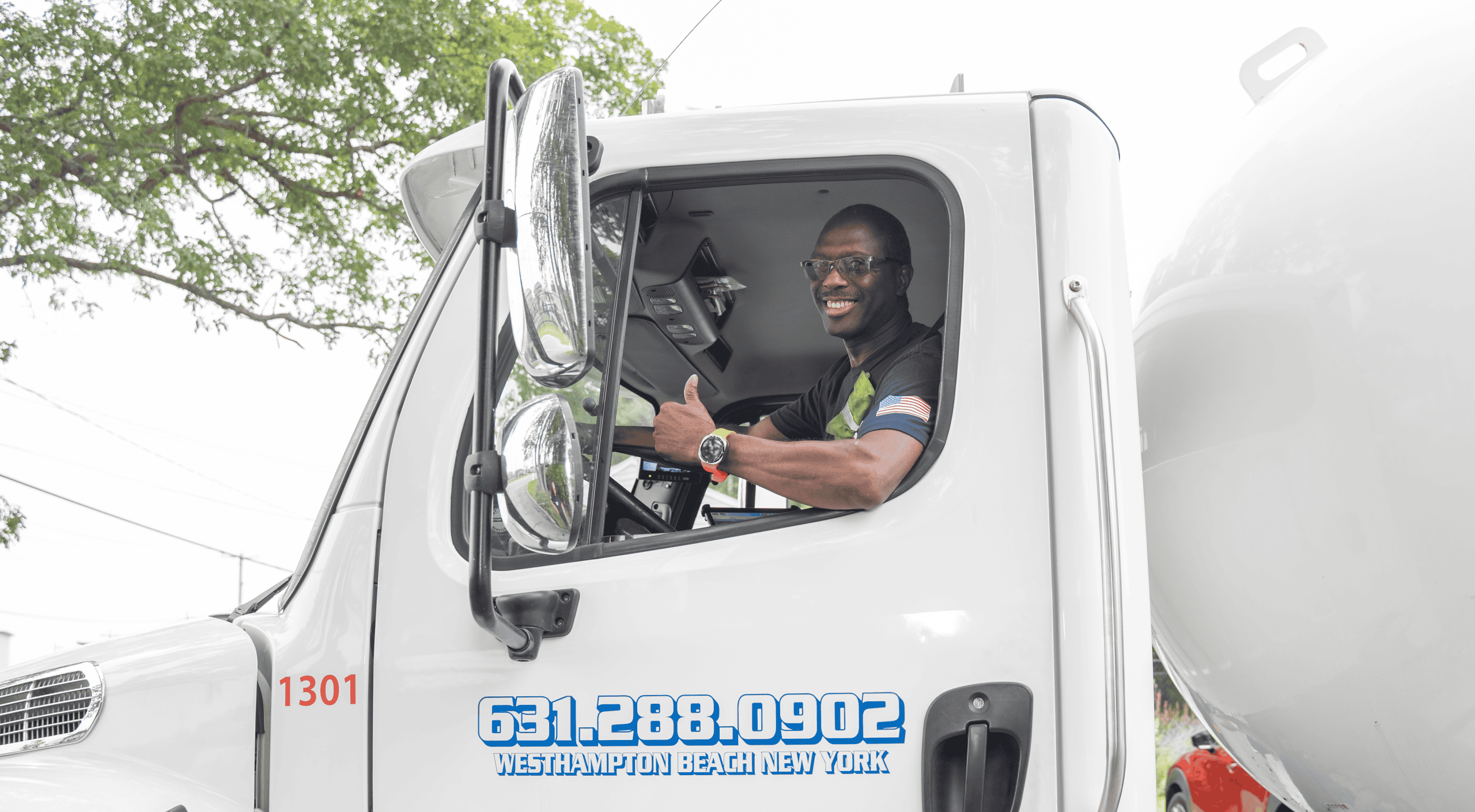 A smiling man gives a thumbs-up from the driver’s seat of a white truck offering Residential Propane Services. The truck has "631.288.0902 Westhampton Beach New York" printed on the door. Trees and power lines are visible outside.